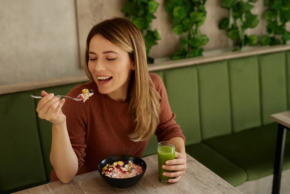 Eine Frau sitzt in einem gemütlichen Café mit grüner Sitzbank und isst lächelnd eine Schüssel Müsli mit frischem Obst. In ihrer anderen Hand hält sie ein Glas grünen Saft – vermutlich ein gesunder Smoothie oder Gemüsesaft.