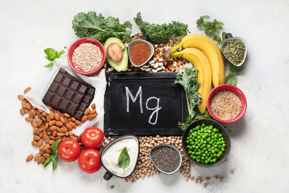 Eine Auswahl magnesiumreicher Lebensmittel ist dekorativ um eine kleine Tafel mit der Aufschrift „Mg“ angeordnet. Zu sehen sind Bananen, Mandeln, dunkle Schokolade, Tomaten, Haferflocken, Avocado, Chiasamen, Joghurt, grüne Erbsen, Vollkornreis und verschiedene grüne Gemüse- und Hülsenfrüchtesorten.