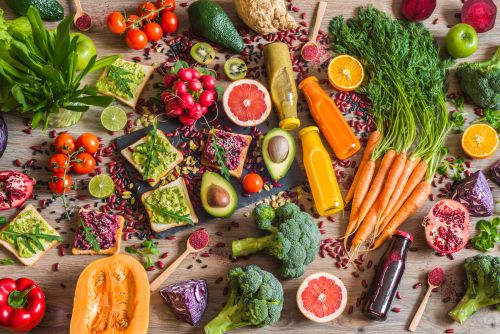Eine bunte Auswahl an frischem Obst, Gemüse und gesunden Lebensmitteln auf einem Holztisch. Zu sehen sind unter anderem Avocados, Brokkoli, Karotten mit Grün, Tomaten, Radieschen, Kiwi, Grapefruits, Orangen, Limetten, Paprika, Kürbis, Rotkohl, Salatblätter, Sprossen, Pomegranate, verschiedene Smoothies in Flaschen sowie Toasts mit Avocado- oder Rote-Bete-Aufstrich, garniert mit Kernen und frischem Grün. Die Szene ist farbenfroh und vermittelt gesunde, vegetarische Ernährung.