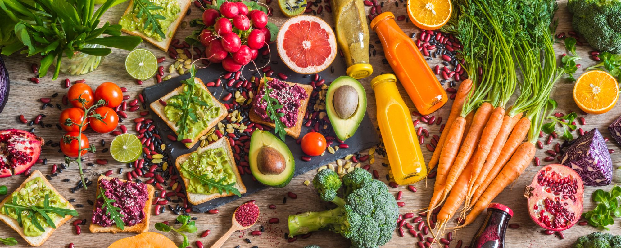Eine bunte Auswahl an frischem Obst, Gemüse und gesunden Lebensmitteln auf einem Holztisch. Zu sehen sind unter anderem Avocados, Brokkoli, Karotten mit Grün, Tomaten, Radieschen, Kiwi, Grapefruits, Orangen, Limetten, Paprika, Kürbis, Rotkohl, Salatblätter, Sprossen, Pomegranate, verschiedene Smoothies in Flaschen sowie Toasts mit Avocado- oder Rote-Bete-Aufstrich, garniert mit Kernen und frischem Grün. Die Szene ist farbenfroh und vermittelt gesunde, vegetarische Ernährung.