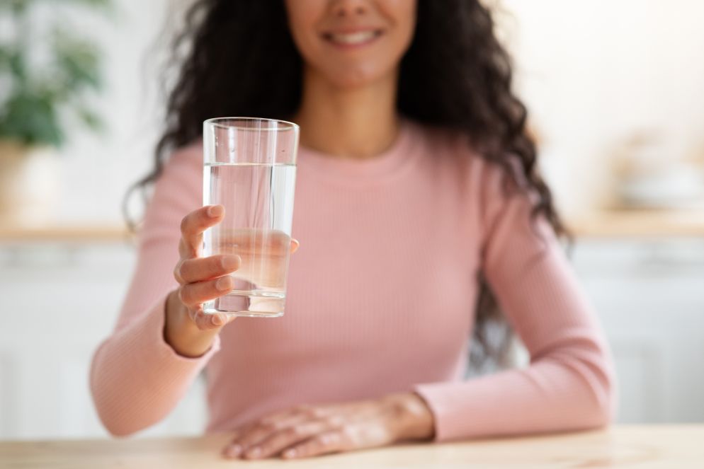 Eine Frau in einem rosa Pullover sitzt an einem Tisch und hält lächelnd ein Glas Wasser in die Kamera. Der Fokus liegt auf dem Glas, das sie mit ausgestreckter Hand präsentiert.