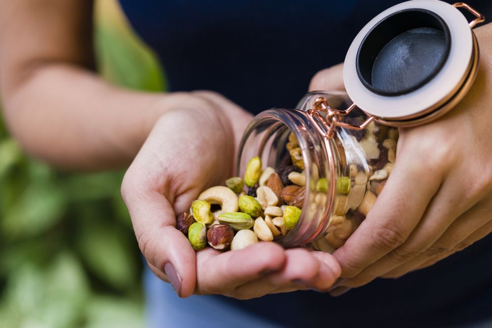 Eine Person hält eine Handvoll gemischter Nüsse, darunter Pistazien, Cashews und Mandeln, die aus einem Glasgefäß in die Hand geschüttet werden. Im Hintergrund sind unscharf grüne Pflanzen zu sehen.