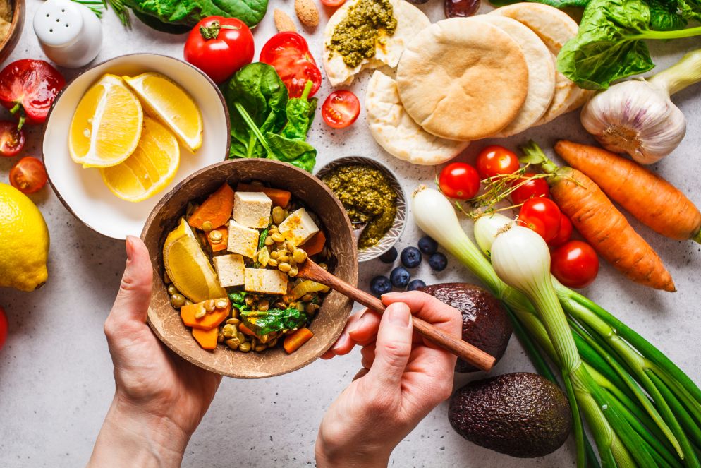 Eine Schüssel mit einer bunten vegetarischen Mahlzeit aus Tofu, Linsen, Spinat, Karotten und einer Zitronenscheibe wird mit beiden Händen gehalten. Um die Schüssel herum liegen frische Zutaten wie Avocados, Tomaten, Karotten, Zwiebeln, Spinat, Pita-Brot und Zitronenscheiben.