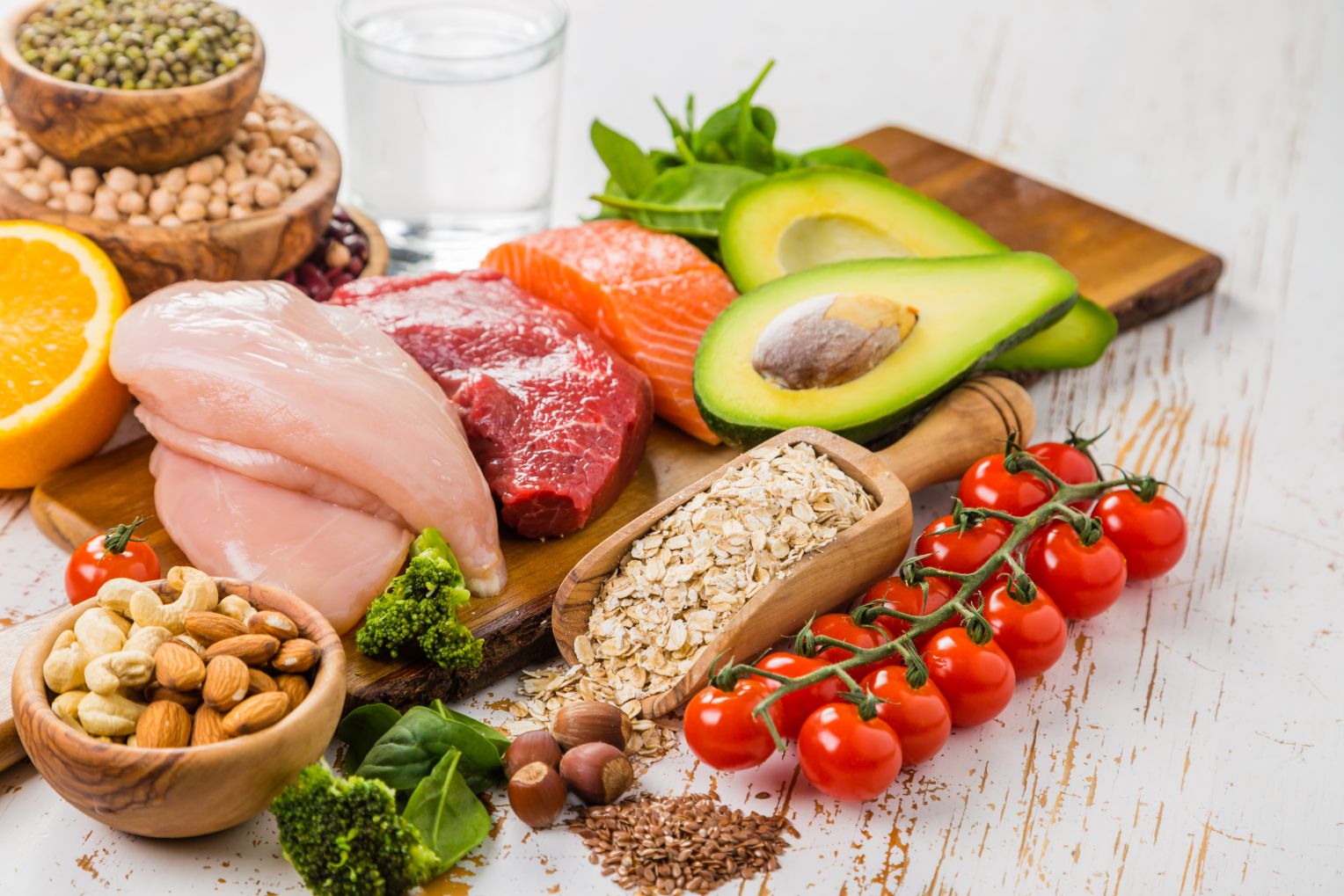 Eine Auswahl gesunder Lebensmittel auf einem Holztisch, bestehend aus rohem Hähnchenfilet, Rindfleisch, Lachs, Avocados, Tomaten, Haferflocken, Nüssen, Brokkoli, Orangen, Hülsenfrüchten und einem Glas Wasser im Hintergrund.