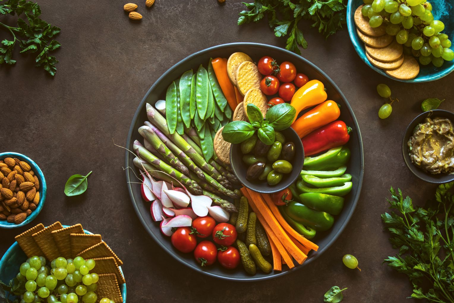 Eine große Servierplatte mit rohem Gemüse wie Paprika, Tomaten, Karotten, Radieschen, Spargel, Zuckerschoten, Oliven und Crackern. Um die Platte herum befinden sich Schalen mit Nüssen, Guacamole und Weintrauben.