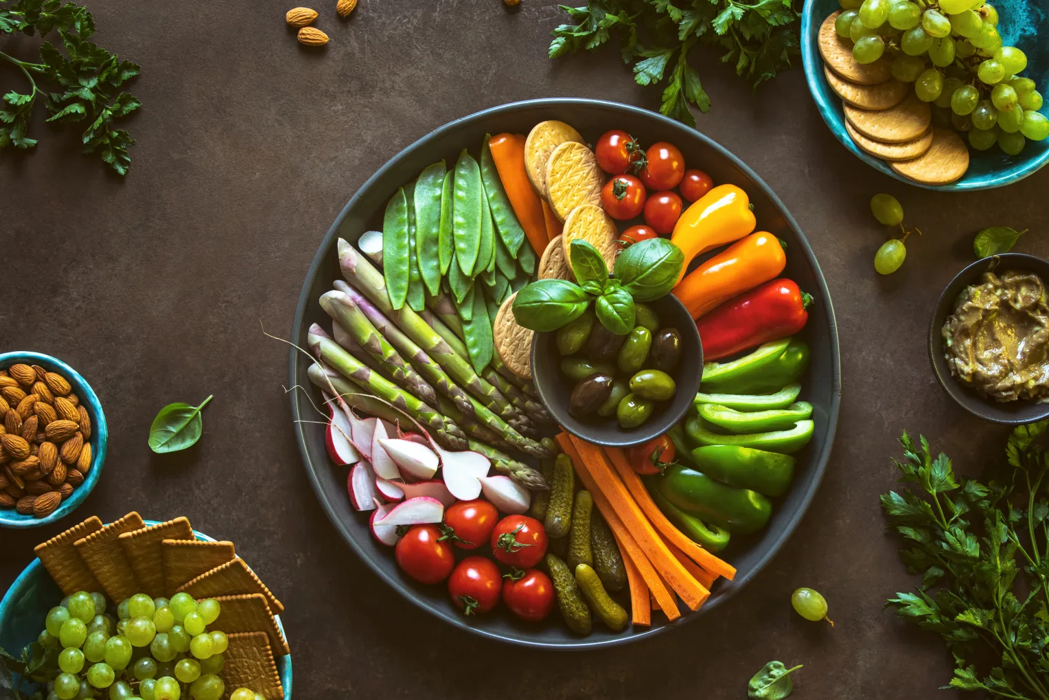 Eine große Servierplatte mit rohem Gemüse wie Paprika, Tomaten, Karotten, Radieschen, Spargel, Zuckerschoten, Oliven und Crackern. Um die Platte herum befinden sich Schalen mit Nüssen, Guacamole und Weintrauben.