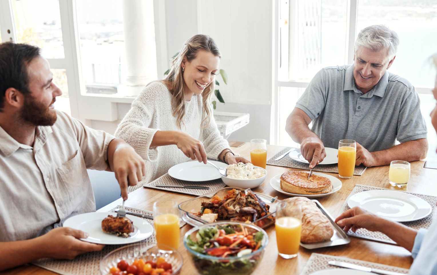 Eine fröhliche Gruppe von Menschen sitzt gemeinsam an einem Esstisch, genießt eine Mahlzeit mit verschiedenen Speisen wie Auflauf, Salat, Quiche, Brot und Gläsern Orangensaft in heller, sonnendurchfluteter Umgebung.