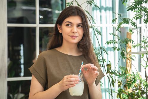 Eine junge Frau steht lächelnd in einem hellen Raum mit Pflanzen im Hintergrund und hält ein Glas mit einem hellen Getränk und einem blauen Strohhalm in der Hand.