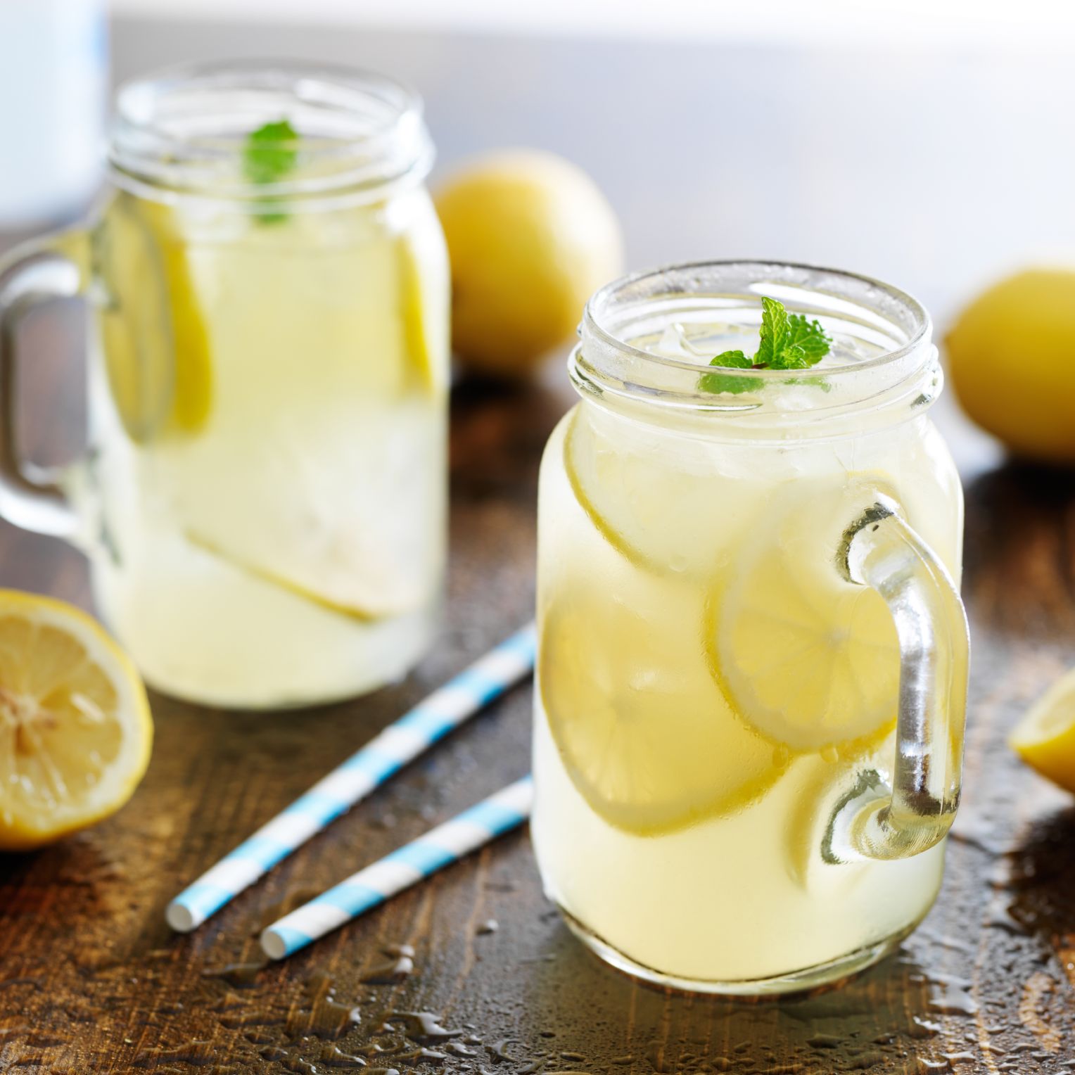 Zwei Gläser mit selbstgemachter Zitronenlimonade mit Eiswürfeln, Zitronenscheiben und Minze auf einem Holztisch, daneben blaue Papierstrohhalme und frische Zitronen.