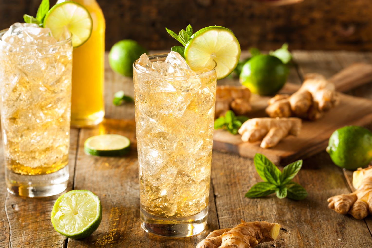 Zwei Gläser mit eisgekühlter Ingwerlimonade, garniert mit Limettenscheiben und Minzblättern, auf einem rustikalen Holztisch mit frischem Ingwer und Limetten.