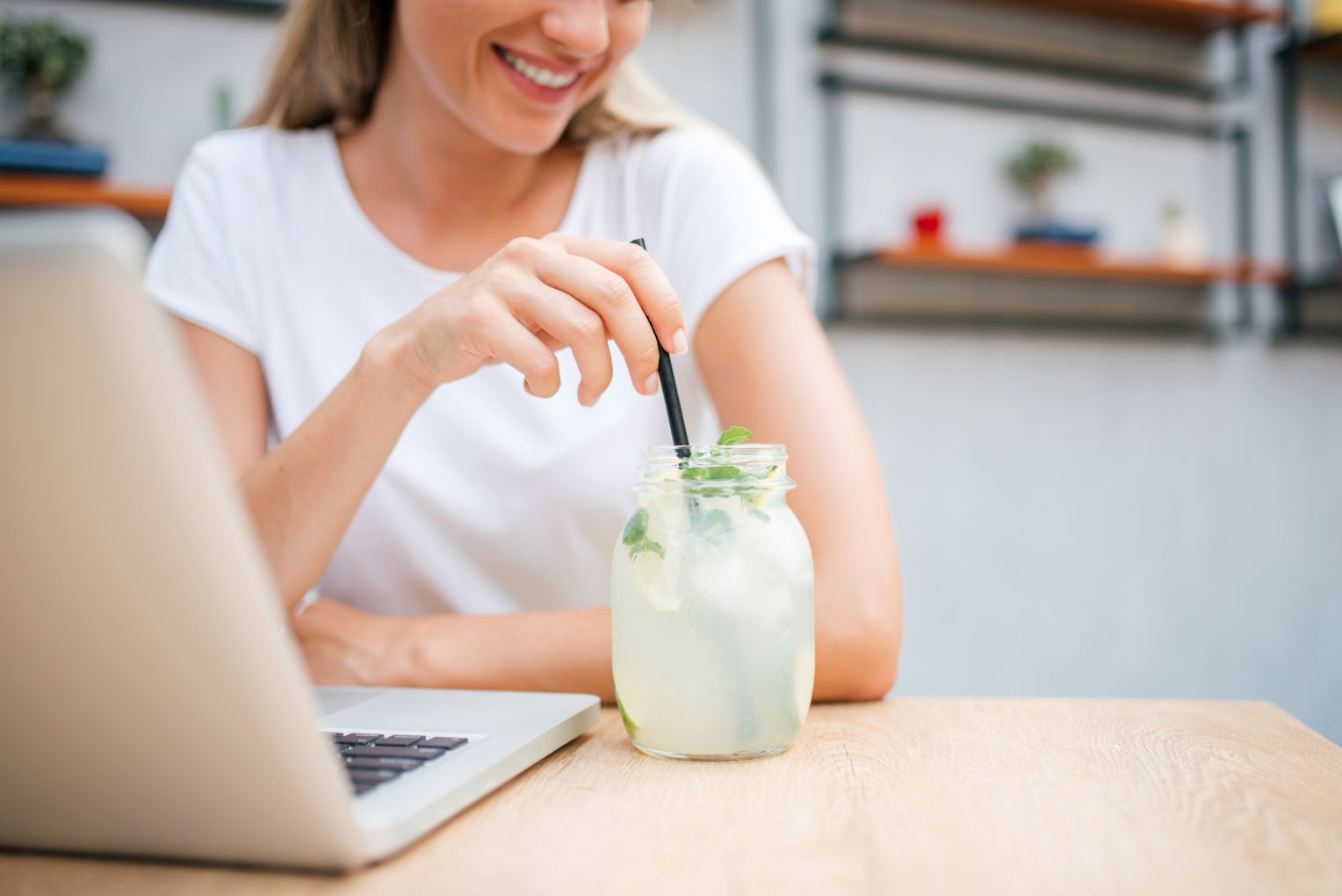 Eine Frau in einem weißen T-Shirt sitzt an einem Tisch mit einem Laptop und rührt mit einem schwarzen Strohhalm in einem Glas Zitronenlimonade mit Minze.