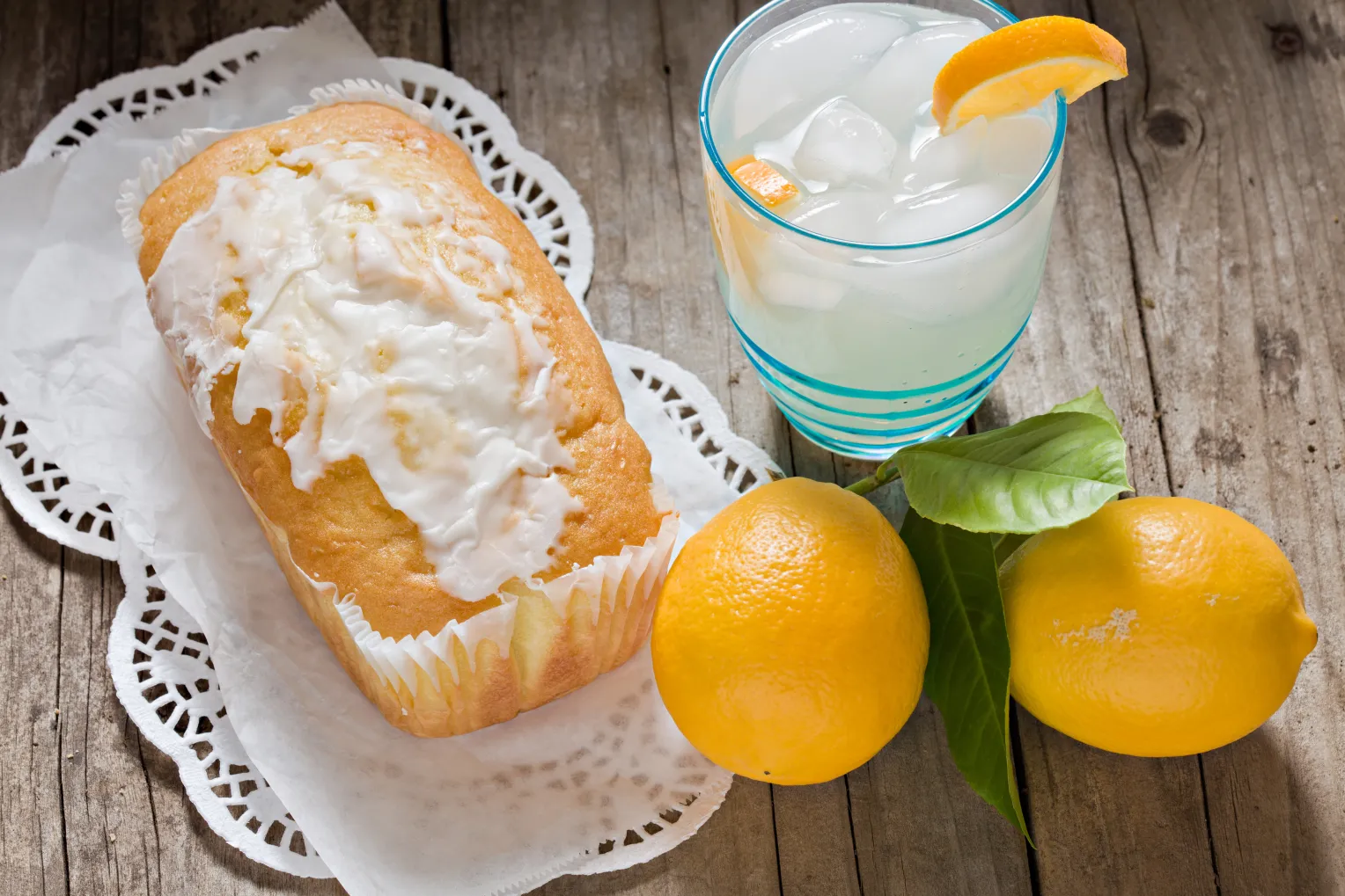 Ein Zitronenkuchen mit Zuckerguss auf einem Spitzenpapier, daneben ein Glas Zitronenlimonade mit Eiswürfeln und Orangenscheibe sowie zwei frische Zitronen.