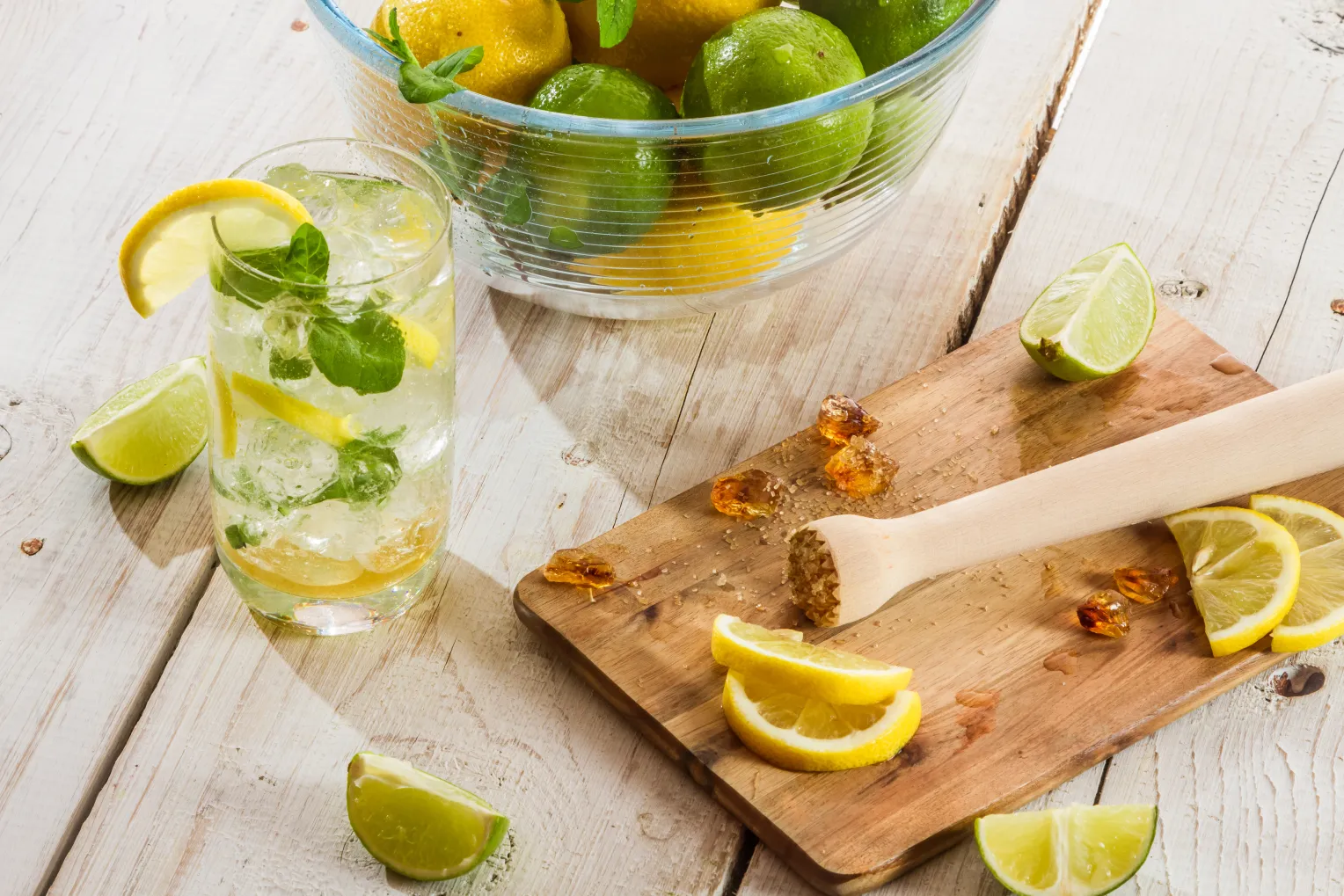 Ein Glas Zitronen-Minz-Getränk mit Eiswürfeln und Zitronenscheiben steht neben einem Schneidebrett mit einem Holzstößel, Zitronenspalten, Kandiszucker und einer Schüssel mit frischen Zitronen und Limetten.