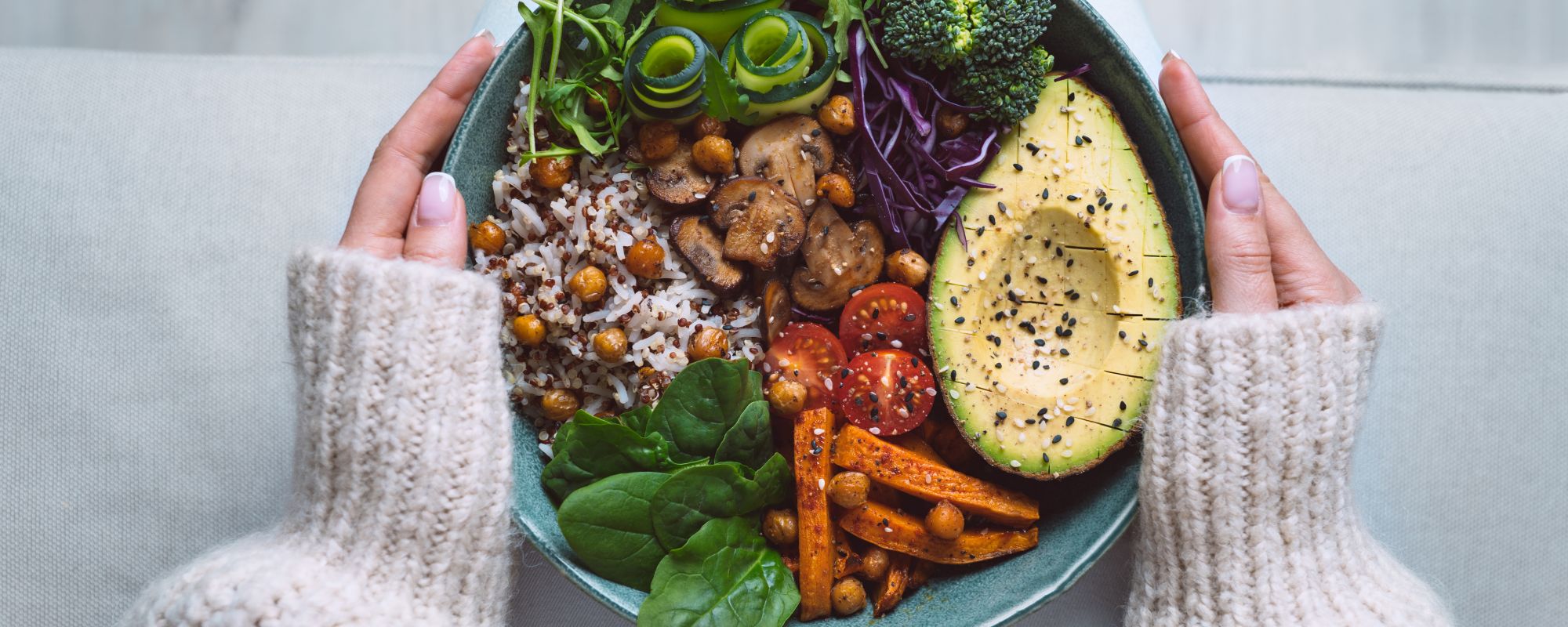 Eine Person in einem gemütlichen, wollenen Pullover hält eine farbenfrohe Buddha-Bowl auf dem Schoß, gefüllt mit Avocado, Brokkoli, rotem Kohl, Kirschtomaten, gebratenen Kichererbsen, Süßkartoffeln, Spinat, Reis-Quinoa-Mix, Zucchini-Röllchen, Rucola und gebratenen Pilzen.