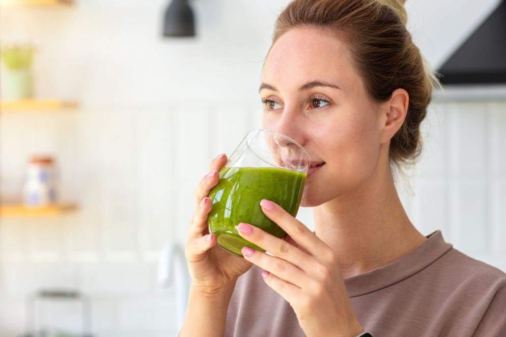 Junge Frau hält ein Glas mit grünem Smoothie in beiden Händen und trinkt in einer hellen Küche.