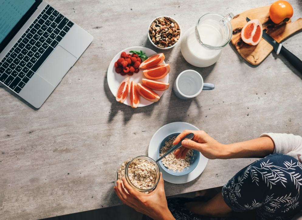 Person bereitet sich an einem Tisch mit Laptop ein Frühstück mit Haferflocken zu, umgeben von Grapefruit, Himbeeren, Nüssen und Milch.