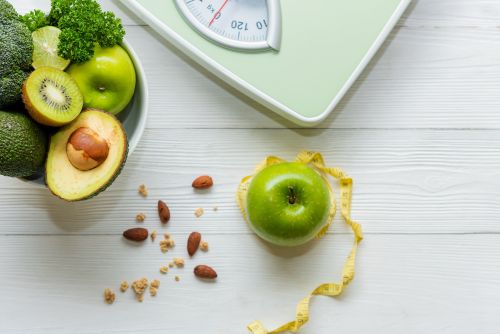 Grüner Apfel mit Maßband auf einem weißen Holztisch, daneben eine Schale mit Avocados, Kiwi, Apfel, Limette und Petersilie sowie eine analoge Personenwaage – Symbol für gesunde Ernährung und Gewichtsmanagement.