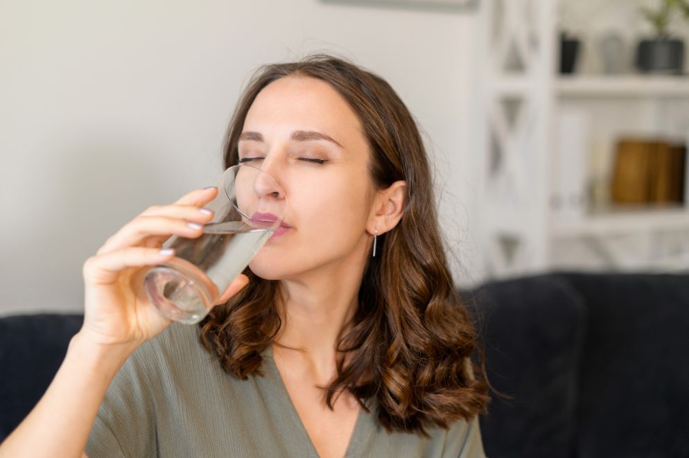 Frau mit geschlossenen Augen trinkt ein Glas klares Wasser, sitzt entspannt auf einem Sofa in einem hellen Wohnzimmer.