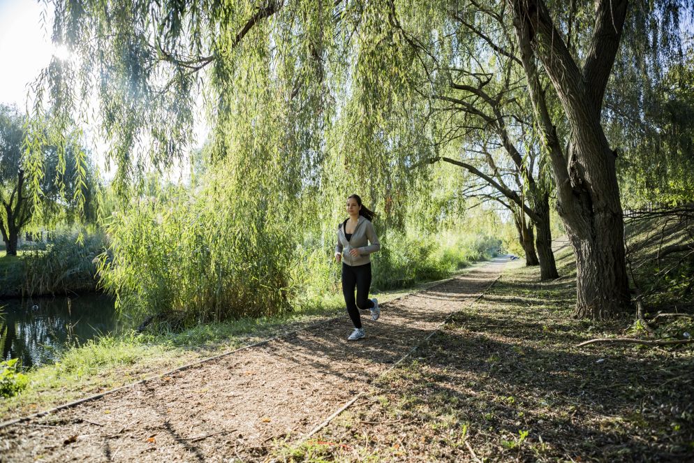 Frau joggt bei Sonnenschein auf einem Naturpfad entlang eines Bachs unter grünen Bäumen im Park.
