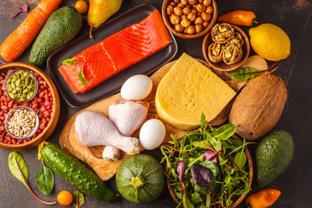 Vielfältige Auswahl an gesunden Low-Carb-Lebensmitteln auf einem dunklen Untergrund, darunter Lachs, Käse, Eier, Hähnchenschenkel, Nüsse, Samen, Avocados, Gemüse und Salat.