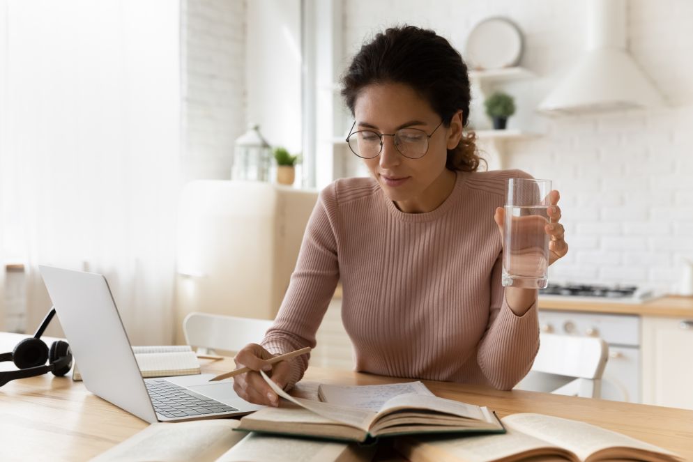 Junge Frau mit Brille sitzt am Schreibtisch, liest ein Buch und hält ein Glas Wasser in der Hand.
