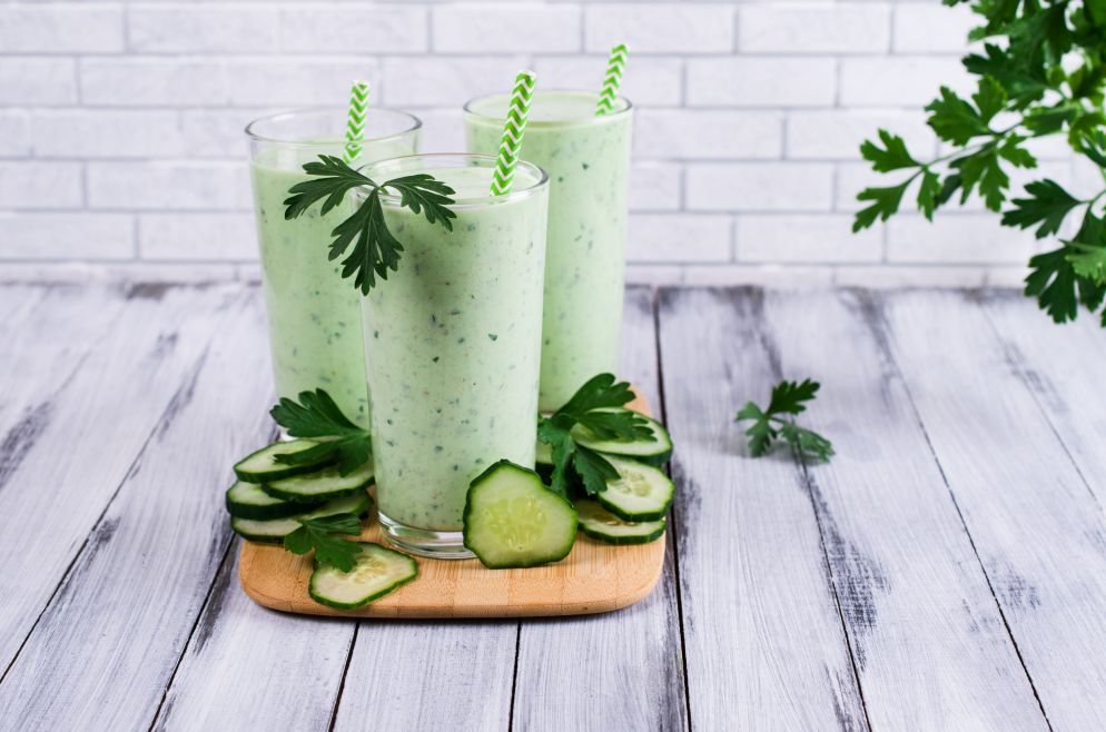 Drei Gläser grüner Smoothie mit Gurke und Petersilie auf einem Holzbrett, dekoriert mit Gurkenscheiben und Kräutern.