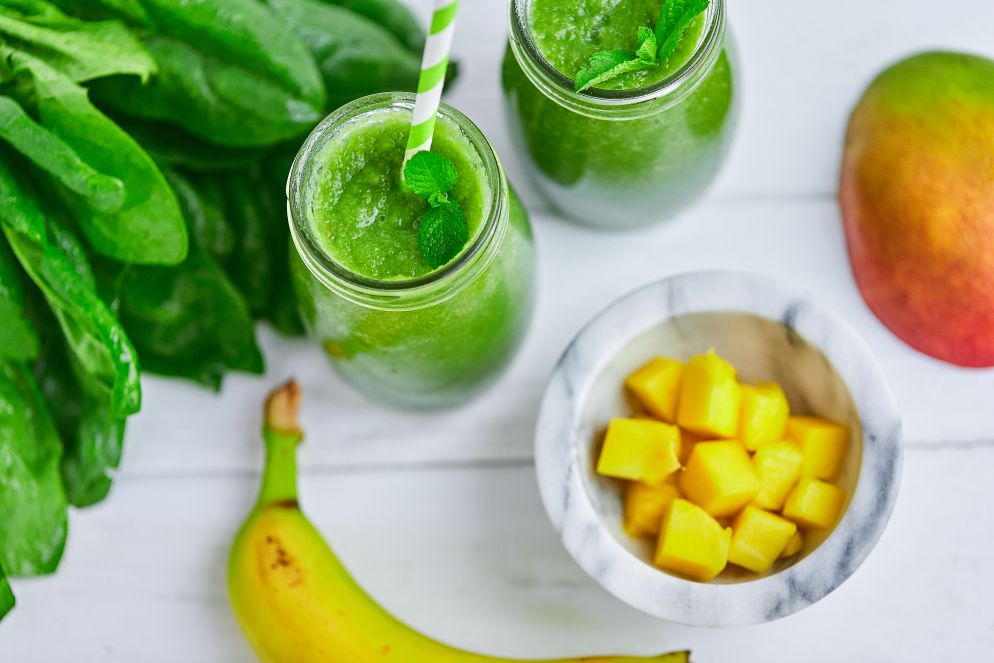 Zwei Gläser grüner Smoothie mit Minze und Strohhalmen, umgeben von Spinatblättern, Banane, Mango und einem Schälchen mit Mangostücken.