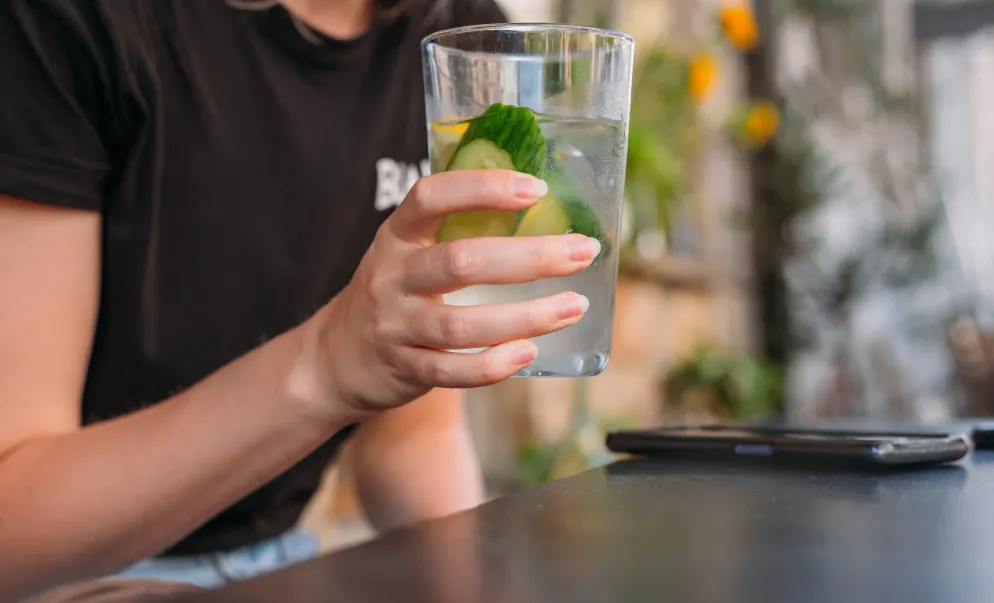 Person hält ein Glas Wasser mit Gurken- und Zitronenscheiben, im Hintergrund unscharfe Pflanzen und ein Tisch.