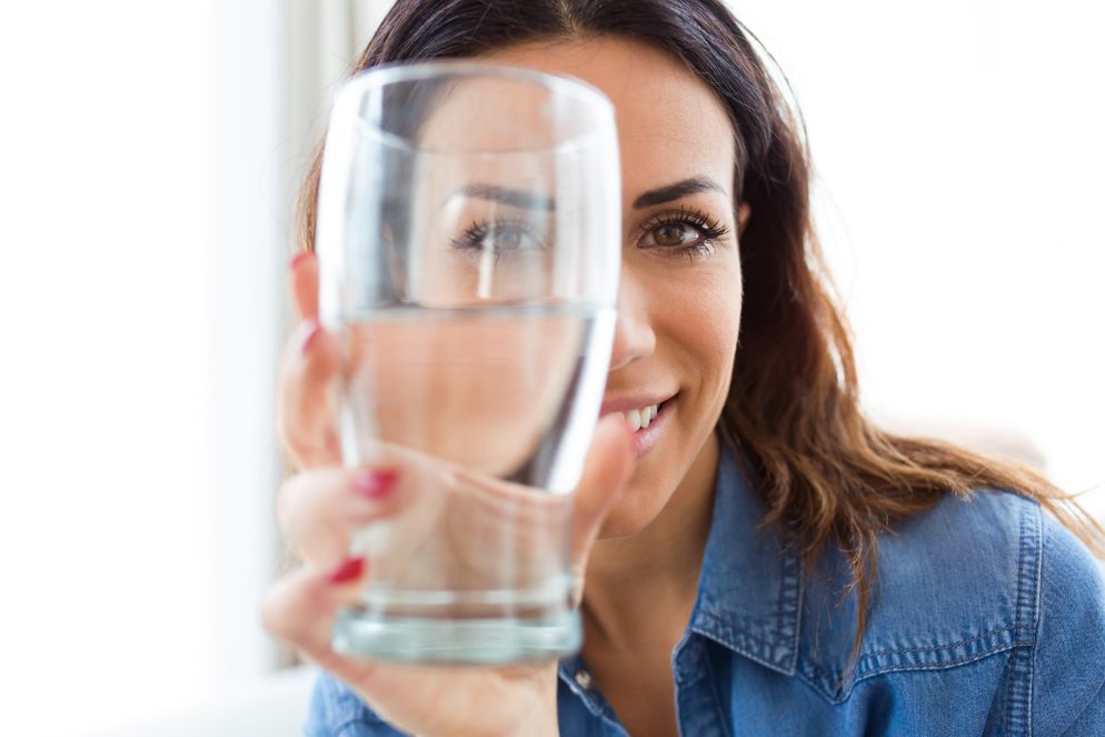 Nahaufnahme einer lächelnden Frau in Jeanshemd, die ein Glas Wasser vor ihr Gesicht hält.