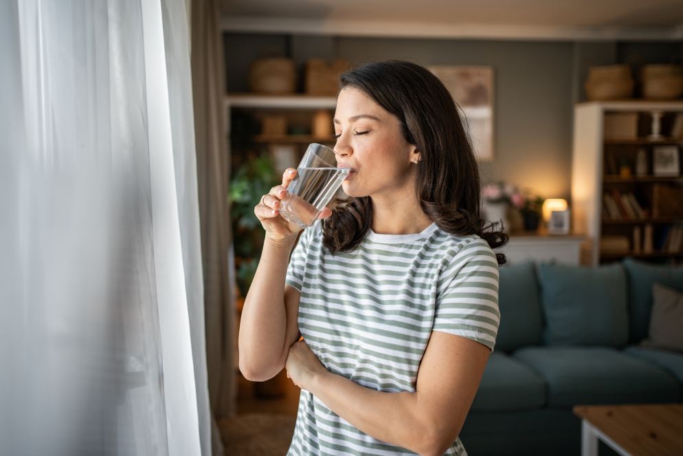 Junge Frau im gestreiften T-Shirt trinkt ein Glas Wasser und schaut entspannt aus dem Fenster in einem gemütlichen Wohnzimmer.