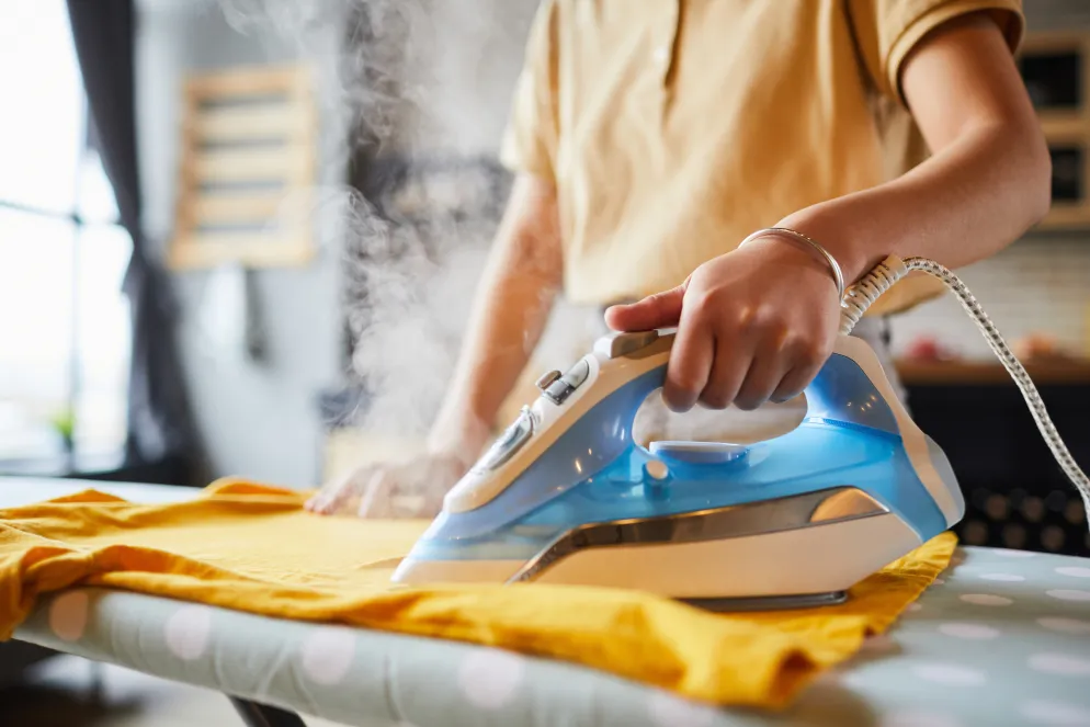 Person bügelt ein gelbes Kleidungsstück mit einem dampfenden Bügeleisen auf einem Bügelbrett.