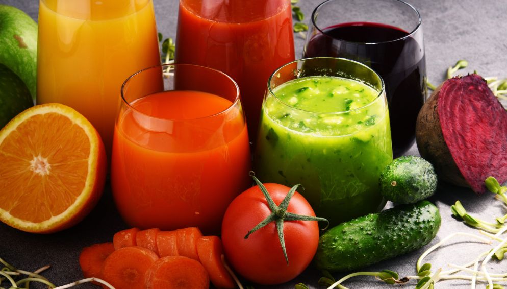Verschiedene bunte Gläser mit Gemüse- und Fruchtsäften, umgeben von frischen Zutaten wie Orange, Tomate, Gurke, Rote Bete und Karotte.