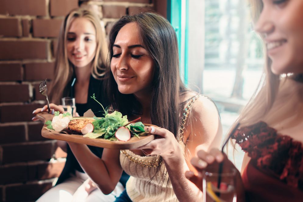 Drei junge Frauen sitzen gemeinsam in einem Restaurant. Eine von ihnen hält einen Holz-Teller mit einem frischen Gericht, das unter anderem Salat und Radieschen enthält, und genießt den Duft mit geschlossenen Augen. Die Stimmung ist fröhlich und entspannt.