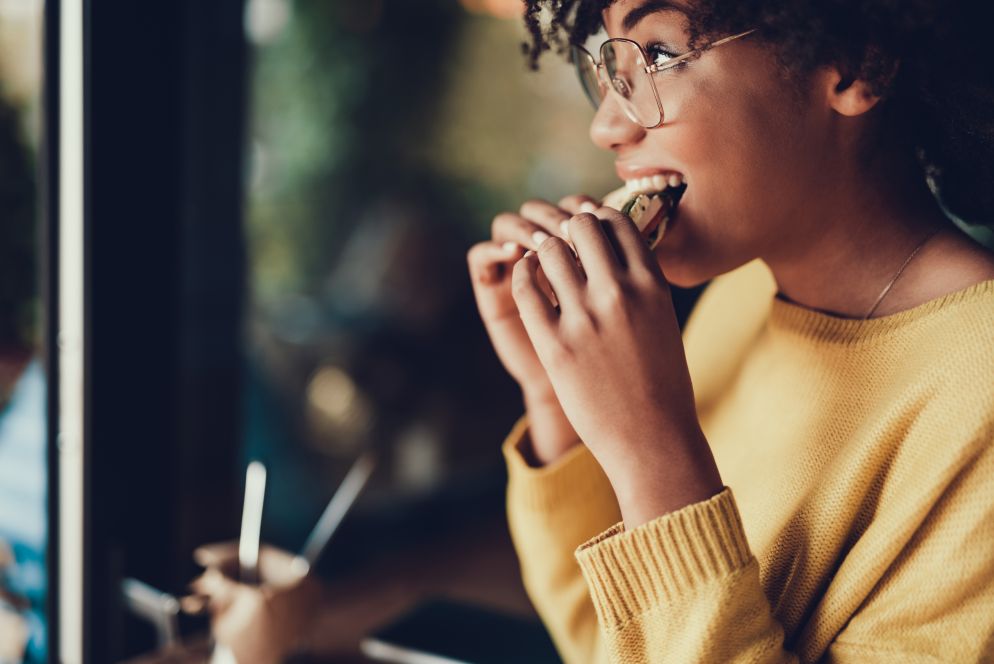 Eine junge Frau mit Brille und gelbem Pullover sitzt am Fenster eines Cafés und beißt genüsslich in einen Burger oder ein Sandwich. Im Hintergrund ist das Café unscharf zu sehen. Sie wirkt zufrieden und entspannt.