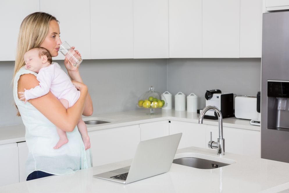 Eine junge Mutter steht in einer modernen weißen Küche, hält ihr schlafendes Baby im Arm und trinkt ein Glas Wasser. Vor ihr steht ein offener Laptop auf der Kücheninsel. Die Szene vermittelt Fürsorge und Alltagsorganisation.