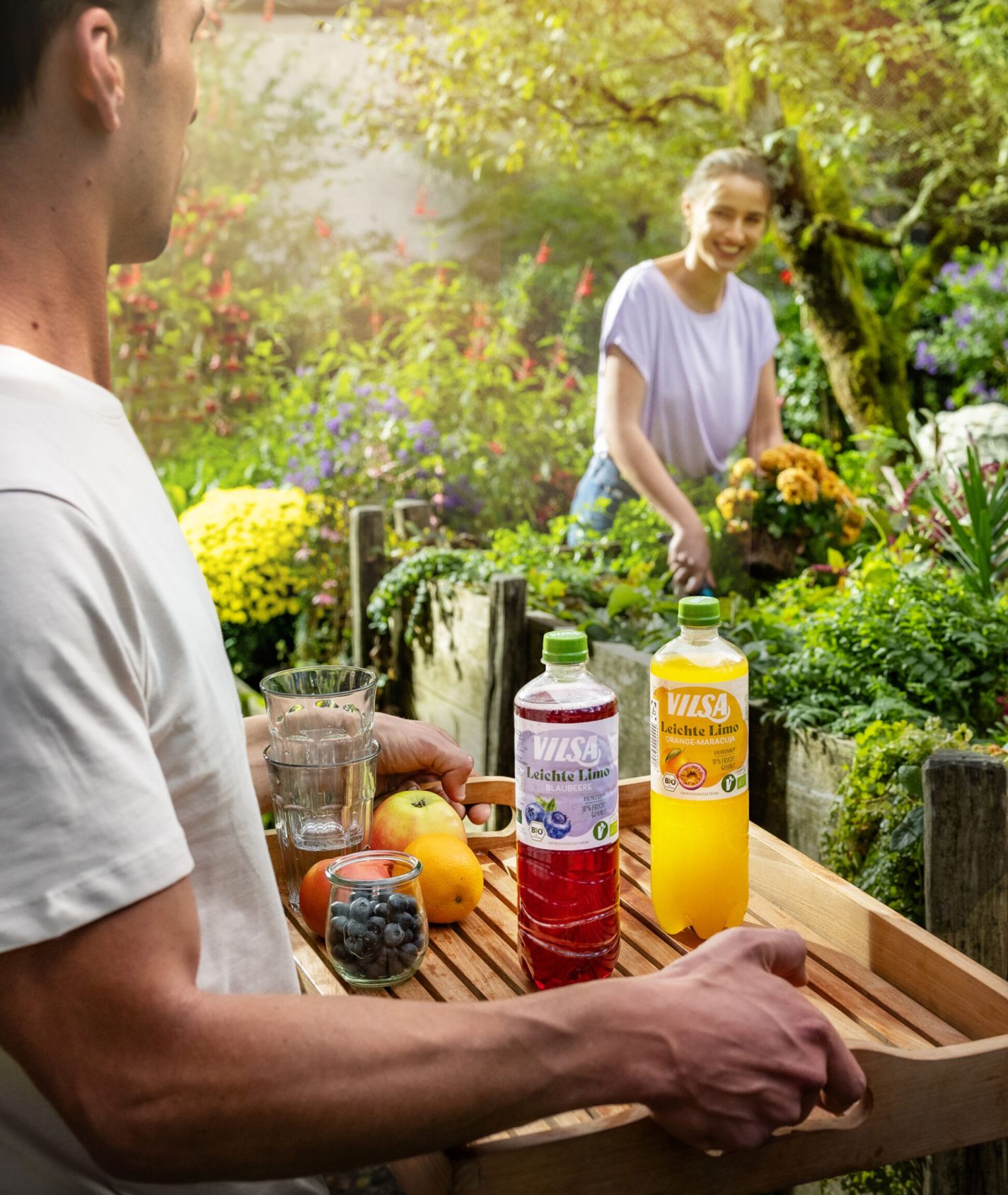 Ein Mann trägt ein Tablett mit Getränken und Obst durch einen farbenfrohen Garten, während eine Frau an einem Hochbeet Blumen pflückt und ihm entgegenlächelt.
