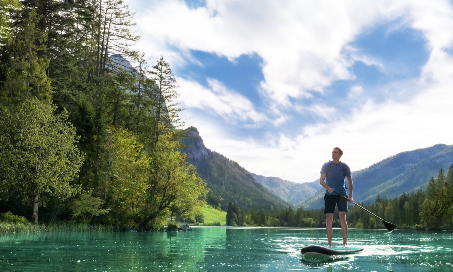Ein Mann steht auf einem Stand-Up-Paddle-Board auf einem glasklaren, türkisfarbenen See, blickt in die Ferne und ist umgeben von bewaldeten Bergen unter einem strahlend blauen Himmel mit leichten Wolken.