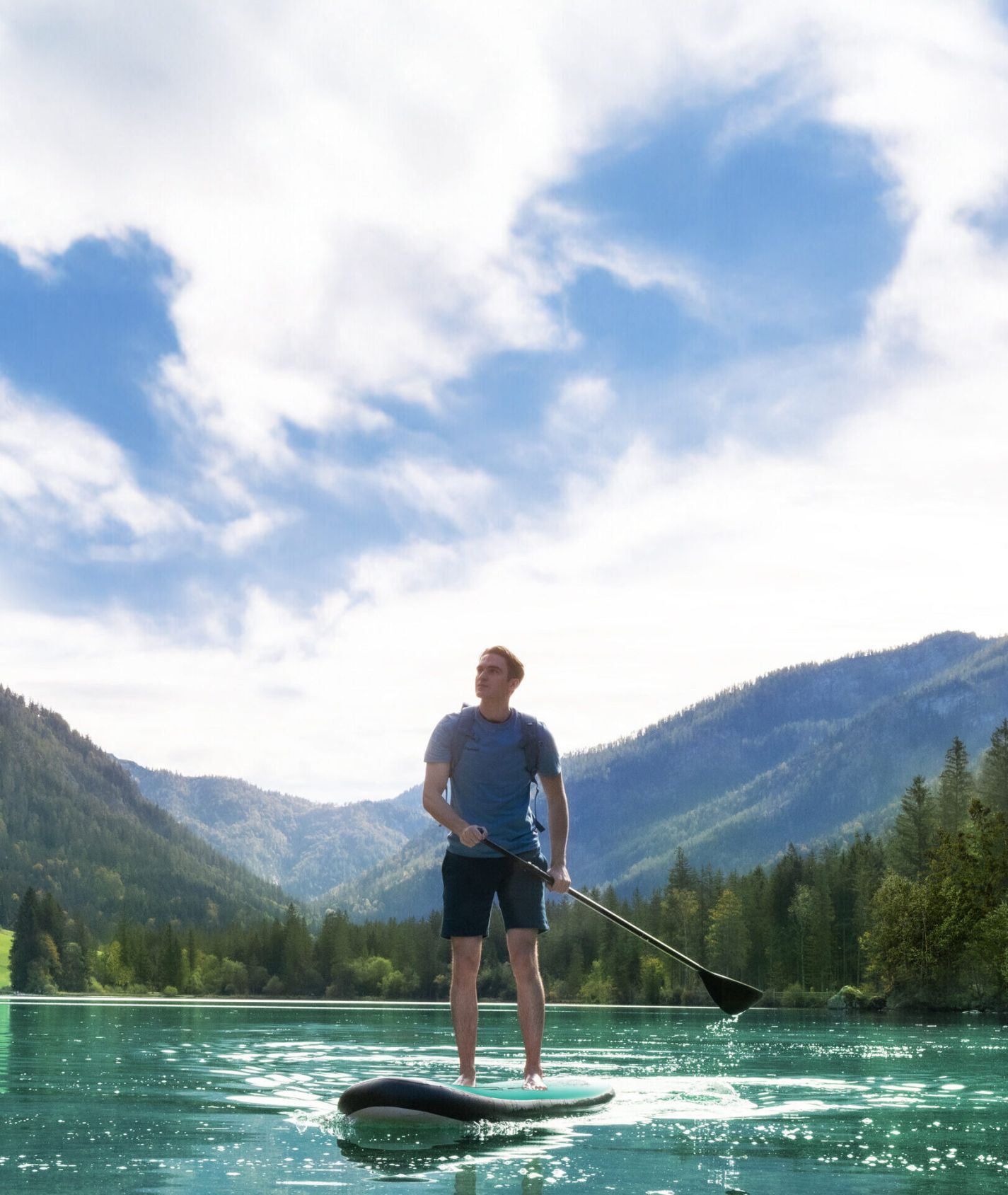Ein Mann steht auf einem Stand-Up-Paddle-Board auf einem glasklaren, türkisfarbenen See, blickt in die Ferne und ist umgeben von bewaldeten Bergen unter einem strahlend blauen Himmel mit leichten Wolken.