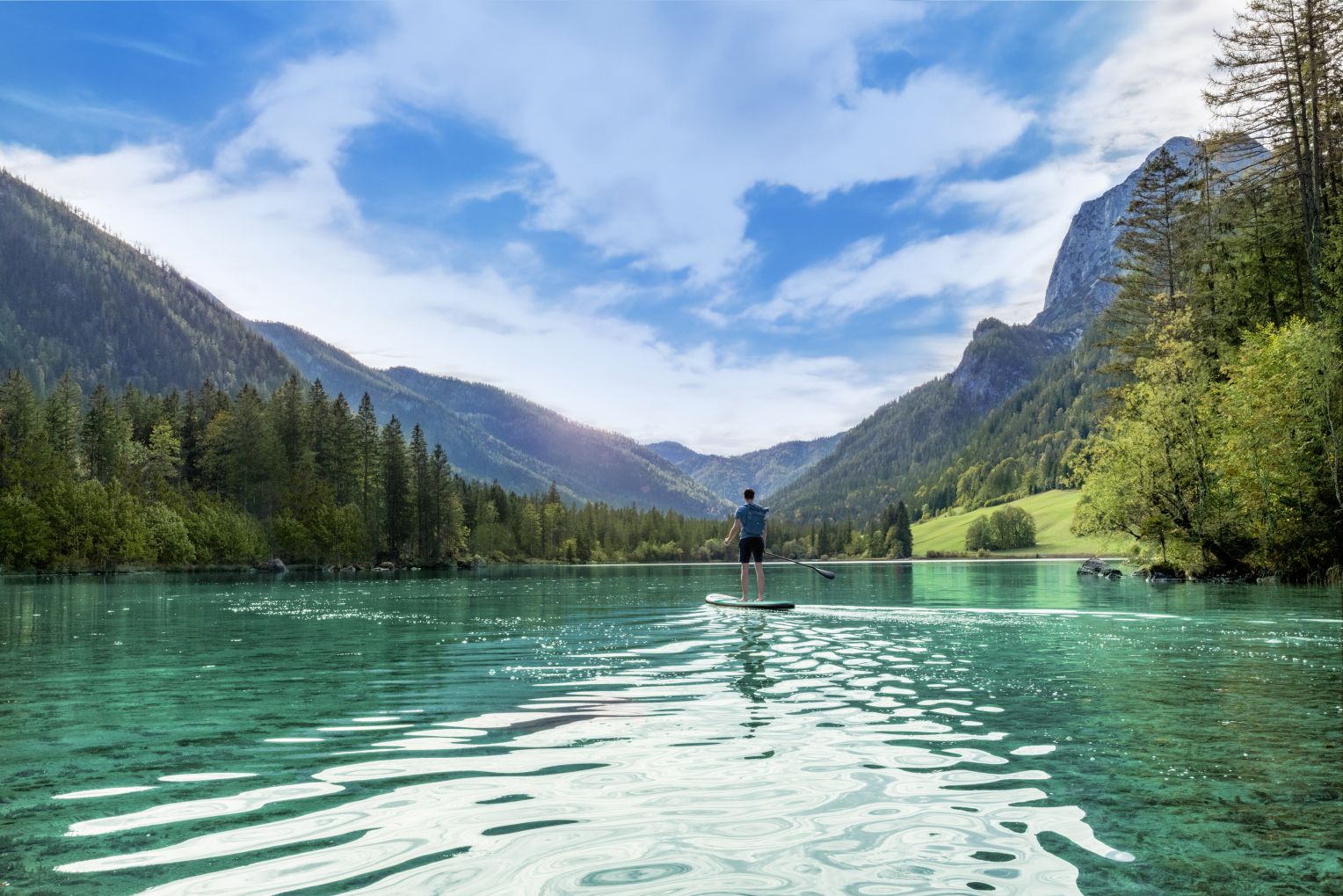 Ein Mann paddelt auf einem Stand-Up-Paddle-Board über einen türkisfarbenen Bergsee, umgeben von dichten Wäldern und hohen Bergen unter blauem Himmel.