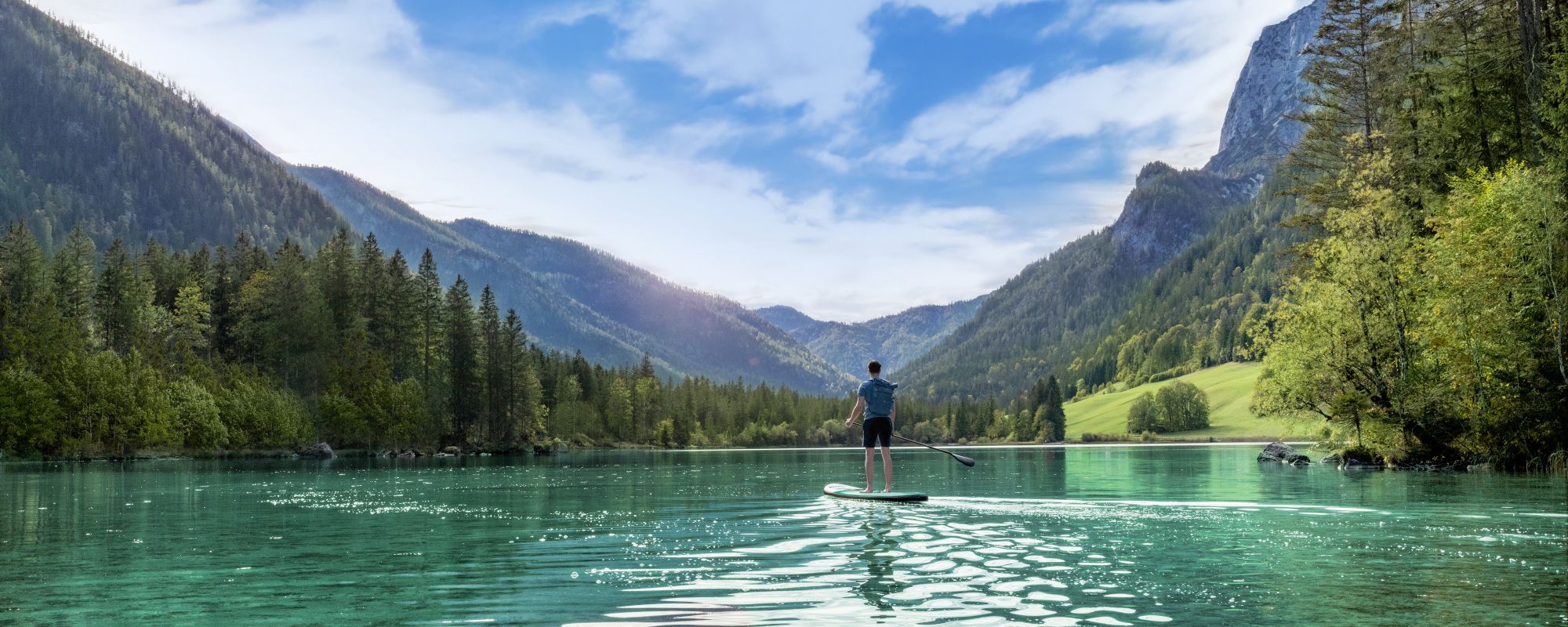 Ein Mann paddelt auf einem Stand-Up-Paddle-Board über einen türkisfarbenen Bergsee, umgeben von dichten Wäldern und hohen Bergen unter blauem Himmel.