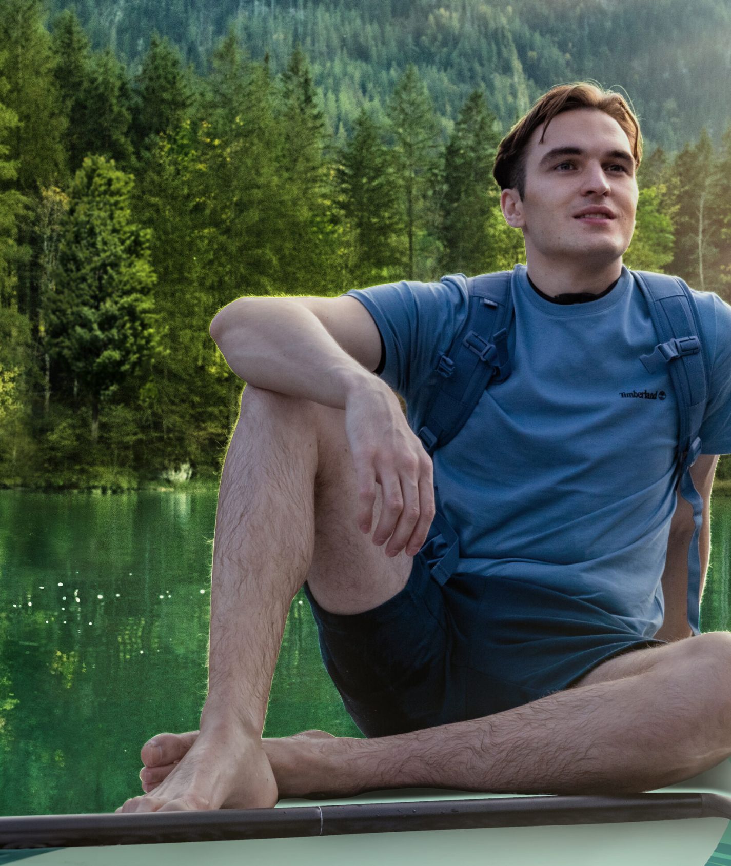 Ein junger Mann sitzt entspannt auf einem Stand-Up-Paddle-Board auf einem smaragdgrünen Bergsee, umgeben von dichtem Wald und Bergen im Sonnenlicht.