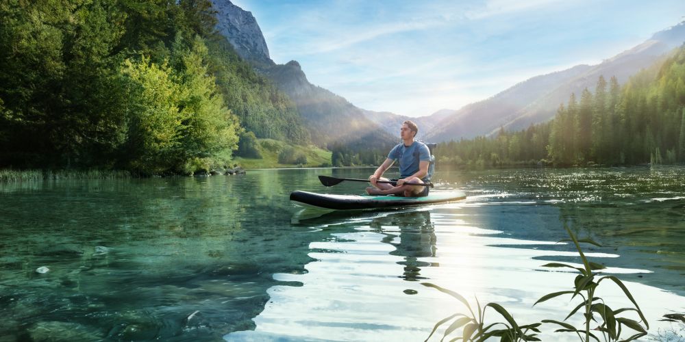 Ein junger Mann sitzt meditierend auf einem Stand-Up-Paddle-Board auf einem klaren, ruhigen Bergsee, umgeben von dicht bewaldeten Hügeln und Bergen im Sonnenlicht.
