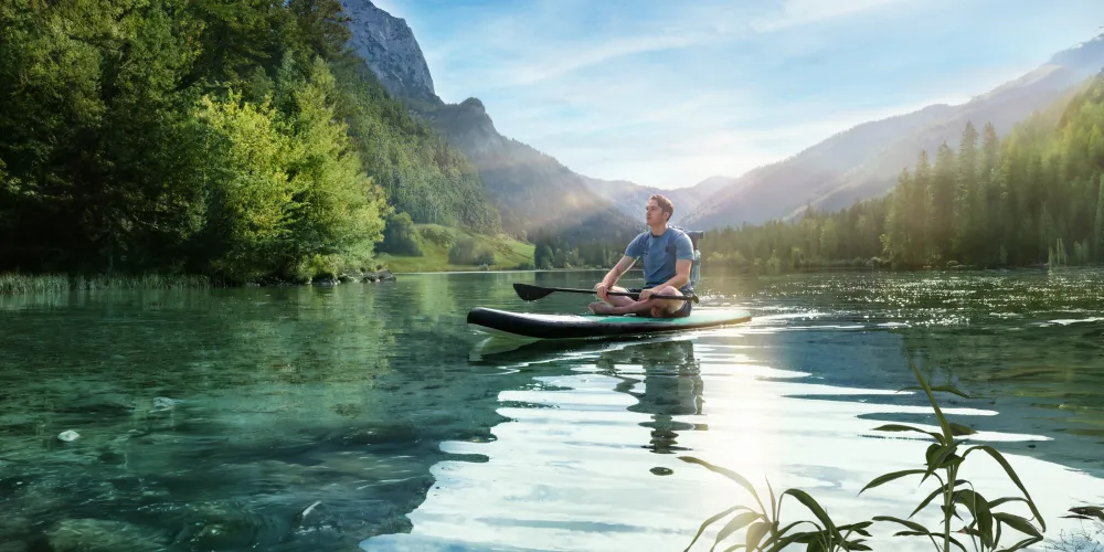 Ein junger Mann sitzt meditierend auf einem Stand-Up-Paddle-Board auf einem klaren, ruhigen Bergsee, umgeben von dicht bewaldeten Hügeln und Bergen im Sonnenlicht.
