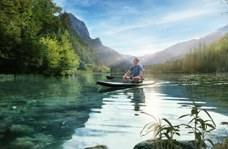 Ein junger Mann sitzt meditierend auf einem Stand-Up-Paddle-Board auf einem klaren, ruhigen Bergsee, umgeben von dicht bewaldeten Hügeln und Bergen im Sonnenlicht.