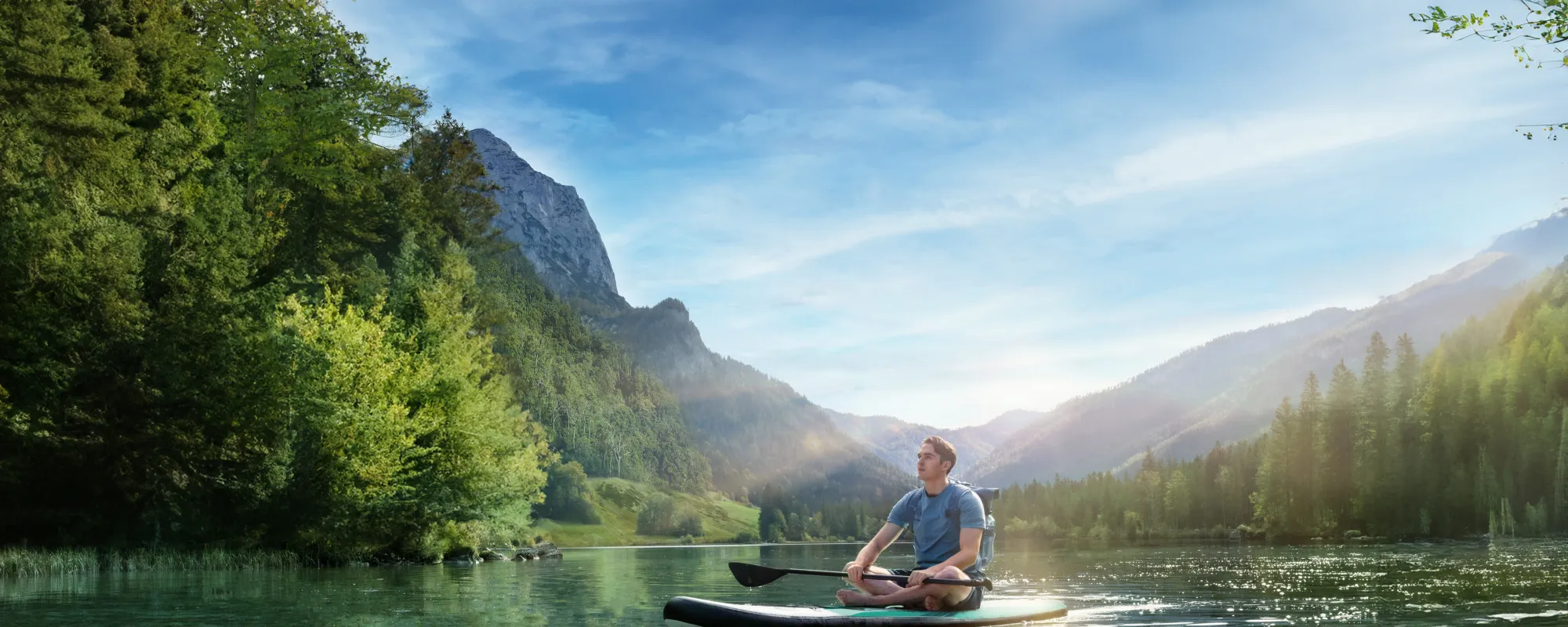 Ein junger Mann sitzt meditierend auf einem Stand-Up-Paddle-Board auf einem klaren, ruhigen Bergsee, umgeben von dicht bewaldeten Hügeln und Bergen im Sonnenlicht.