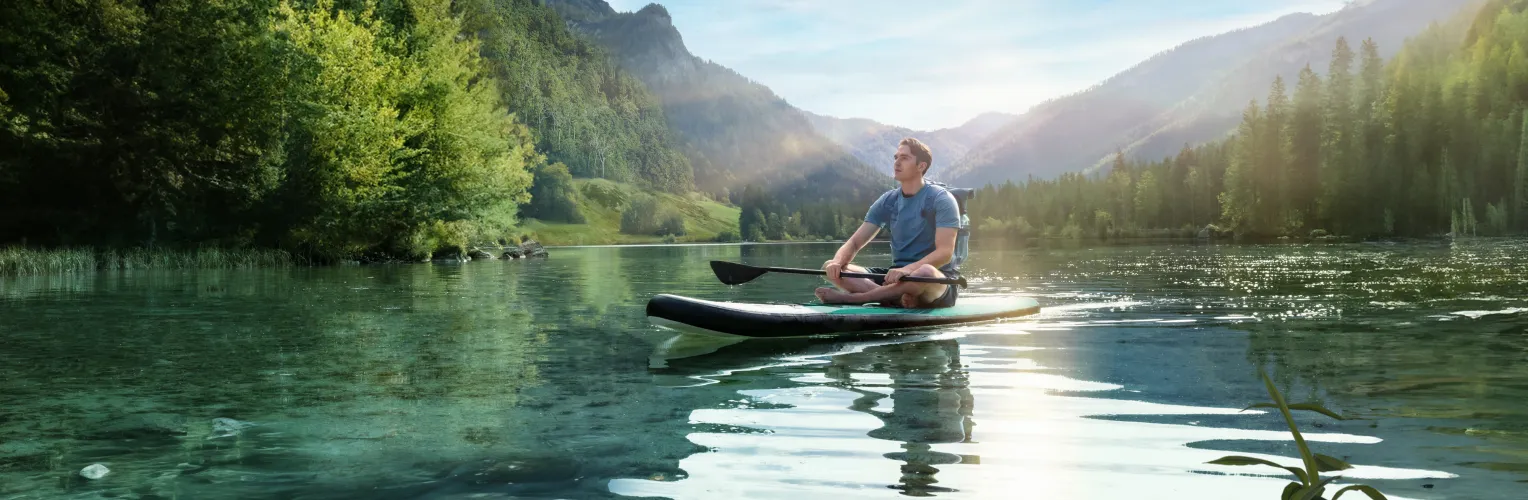 Ein junger Mann sitzt meditierend auf einem Stand-Up-Paddle-Board auf einem klaren, ruhigen Bergsee, umgeben von dicht bewaldeten Hügeln und Bergen im Sonnenlicht.