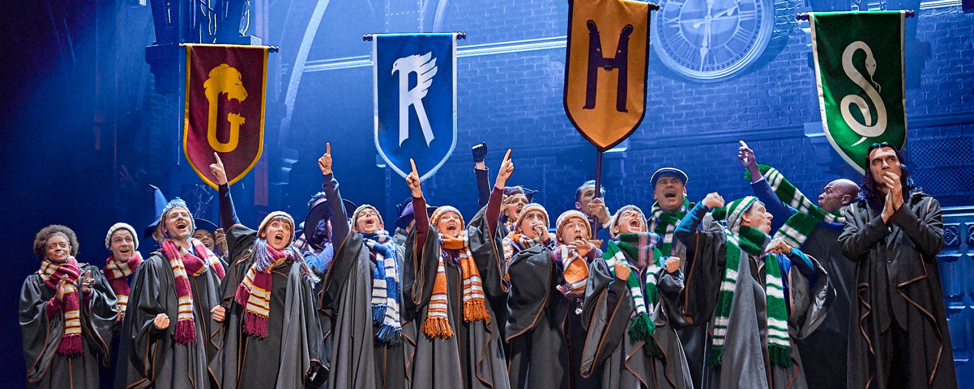 Eine Gruppe von Schauspielerinnen und Schauspielern in Hogwarts-Schuluniformen steht auf einer Bühne, singt oder ruft begeistert und zeigt mit erhobenen Fingern nach oben. Über ihnen hängen vier Fahnen mit den Haus-Symbolen von Hogwarts: Gryffindor (G), Ravenclaw (R), Hufflepuff (H) und Slytherin (S). Die Szene ist Teil einer Theaterproduktion, wahrscheinlich „Harry Potter und das verwunschene Kind“, mit dramatischer Beleuchtung und gotischem Bühnenbild im Hintergrund.