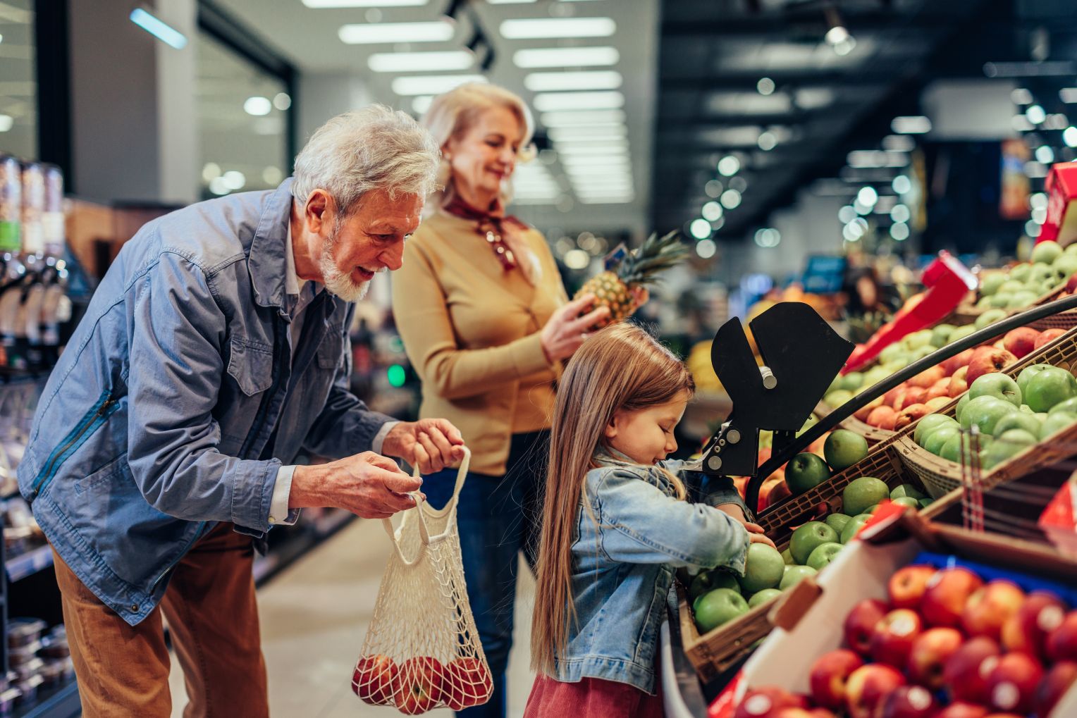 Ein älteres Ehepaar und ein kleines Mädchen kaufen frisches Obst in einem Supermarkt. Der Großvater hält ein Netz voller Äpfel, während das Mädchen grüne Äpfel auswählt.
