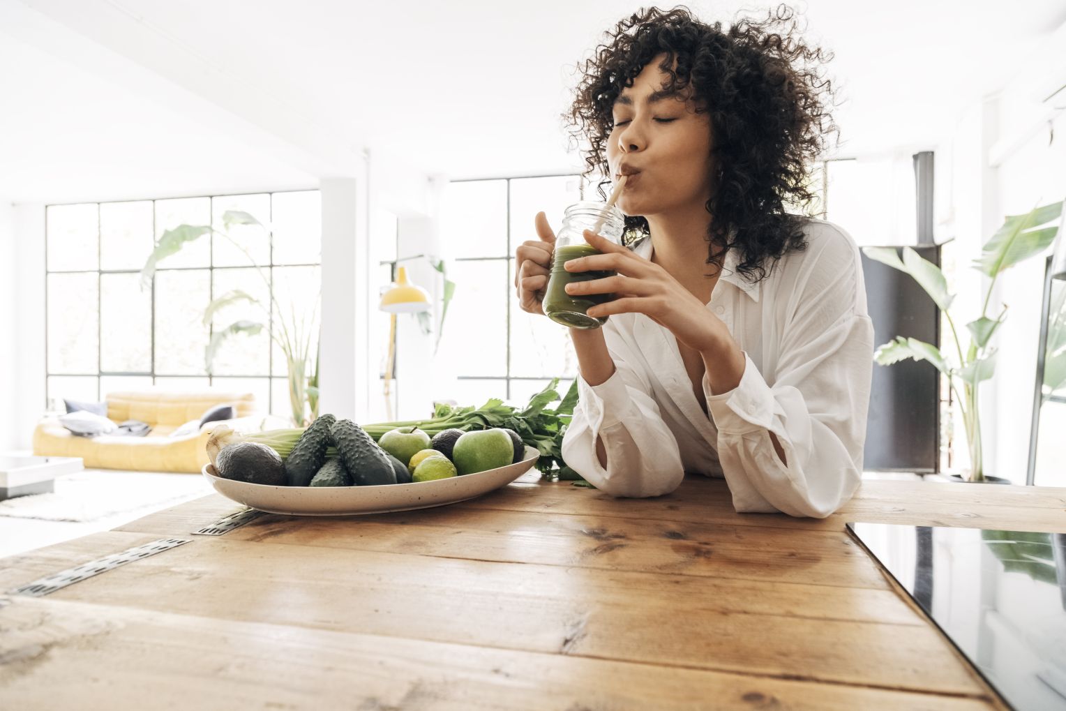 Frau trinkt einen grünen Smoothie an einem Holztisch mit frischem Obst und Gemüse, darunter Avocados, Gurken, Limetten und Sellerie. Der Raum ist hell und modern eingerichtet.