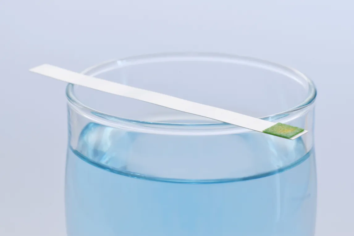 Ein Glas mit bläulichem Wasser und einem pH-Teststreifen, der oben auf dem Glas liegt. Der Teststreifen zeigt eine grün-gelbe Verfärbung an der Spitze.